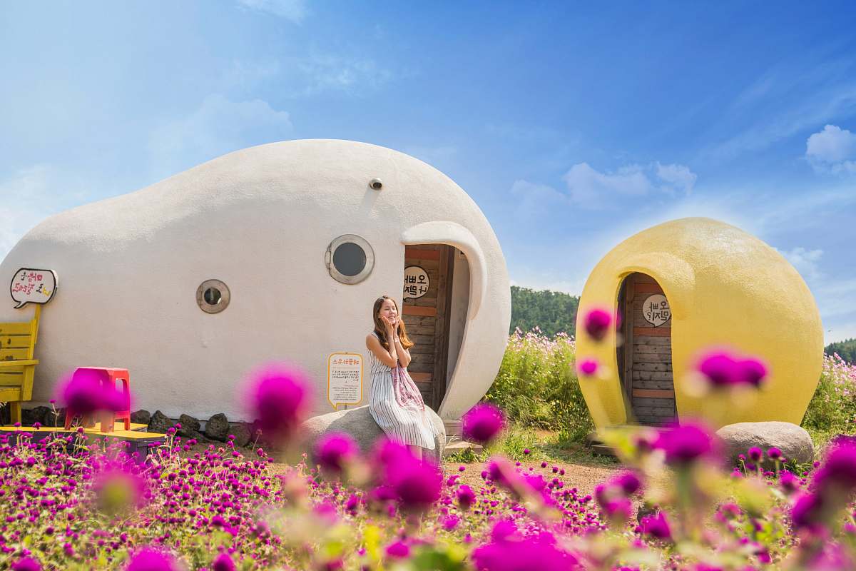 jeju-do woman sitting in front of white and brown rock house surrounded with purple flowers under blue sky figurine
